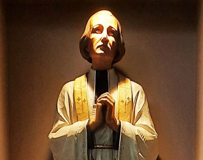 A wooden carving of St. John Vianne robed in white, his hands clasped, and eyes fixed sky-ward.
